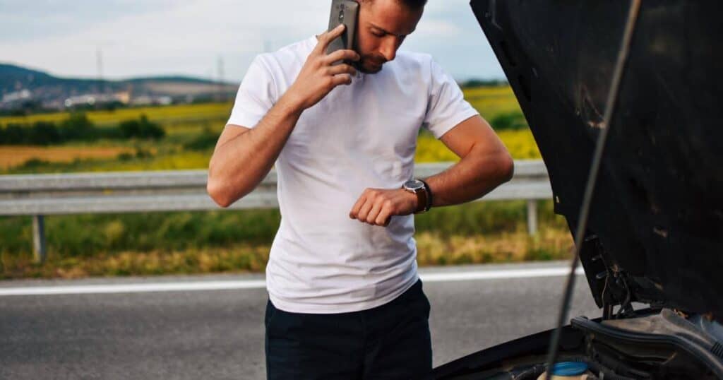 production worker on cell phone and looking at watch, standing on side of road with broken down car